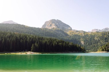 Berrak turkuaz göl yemyeşil ormanlarla ve güneşli bir günde parlak bir gökyüzünün altında dağlarla çevrili.