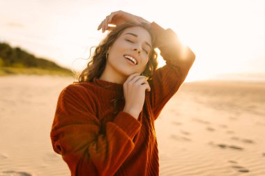 Gün batımında güneşin sıcağının tadını çıkaran genç bir kadın, yumuşak dalgalar ve altın ışıkla neşeli ifadesini güçlendiriyor.