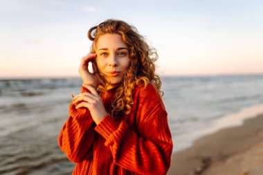 Gün batımında güneşin sıcağının tadını çıkaran genç bir kadın, yumuşak dalgalar ve altın ışıkla neşeli ifadesini güçlendiriyor.