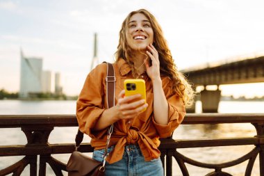 Genç bir kadın, gün batımında deniz kenarında canlı bir şehirde, köprüde akıllı telefonunu kullanırken gülümsüyor.
