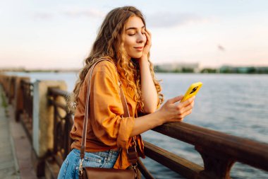 Genç bir kadın, gün batımında deniz kenarında canlı bir şehirde, köprüde akıllı telefonunu kullanırken gülümsüyor.