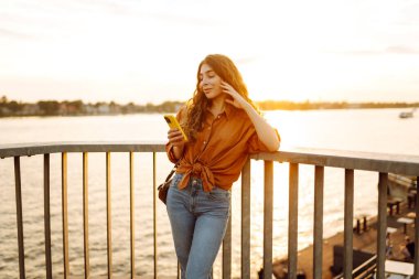 Genç bir kadın, gün batımında deniz kenarında canlı bir şehirde, köprüde akıllı telefonunu kullanırken gülümsüyor.