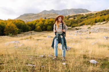 Genç bir kadın güneşli bir öğleden sonra yürüyüşünün tadını çıkarıyor. Manzaralı dağlarda, canlı sonbahar yaprakları arasında kazak ve şapka giyiyor.