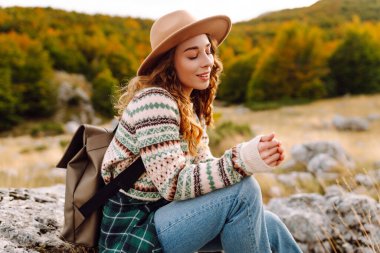Genç bir kadın güneşli bir öğleden sonra yürüyüşünün tadını çıkarıyor. Manzaralı dağlarda, canlı sonbahar yaprakları arasında kazak ve şapka giyiyor.
