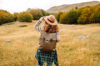 Genç bir kadın güneşli bir öğleden sonra yürüyüşünün tadını çıkarıyor. Manzaralı dağlarda, canlı sonbahar yaprakları arasında kazak ve şapka giyiyor.