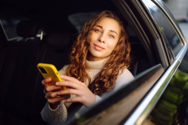 Kıvırcık saçlı genç bir kadın, güneşli bir günde arabada otururken akıllı telefon kullanır. Teknoloji kavramı, internet, blog, arabayla seyahat, iş