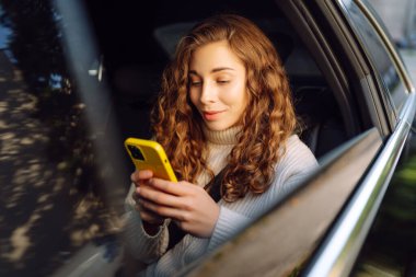 Kıvırcık saçlı genç bir kadın, güneşli bir günde arabada otururken akıllı telefon kullanır. Teknoloji kavramı, internet, blog, arabayla seyahat, iş