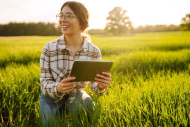 Genç bir kadın yemyeşil çimenlerde diz çökerken tablet kullanarak gülümsüyor. Zirai üretimde elektronik, teknoloji ve mobil uygulamalar kullanarak modern akıllı tarım kavramı.