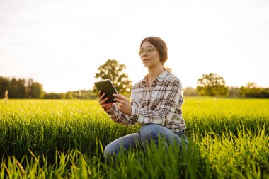 Genç bir kadın yemyeşil çimenlerde diz çökerken tablet kullanarak gülümsüyor. Zirai üretimde elektronik, teknoloji ve mobil uygulamalar kullanarak modern akıllı tarım kavramı.
