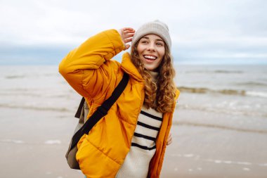 Sarı ceketli genç bir kadın okyanus kıyısındaki sahilde gülümsüyor, gri gökyüzüyle rüzgarlı bir günün tadını çıkarıyor. Sakinlik ve sükunet. Seyahat, turizm konsepti.