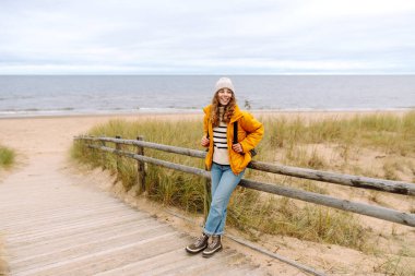 Sarı ceketli genç bir kadın okyanus kıyısındaki sahilde gülümsüyor, gri gökyüzüyle rüzgarlı bir günün tadını çıkarıyor. Sakinlik ve sükunet. Seyahat, turizm konsepti.