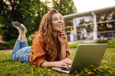 Güneşli bir öğleden sonranın tadını çıkaran genç bir kadın yeşil ve kentsel manzarayla çevrili bir parkta dizüstü bilgisayarıyla çalışıyor. Serbest Çalışan, Çevrimiçi Eğitim.