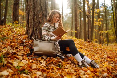 Genç bir kadın renkli yaprakların üzerinde otururken bir kitaba dalmış, sessiz bir anın tadını çıkarıyor. - Evet. Eğitim. Doğadan zevk alma kavramı.