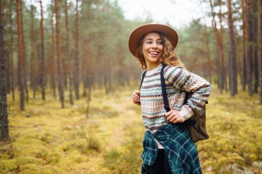 Ormanda şapka ve sırt çantası takan genç bir kadın, sonbaharda uzun ağaçların arasında huzurlu bir yürüyüş yapıyor. Orman banyosu. Etkin yaşam biçimi