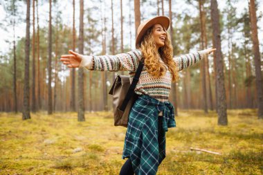 Ormanda şapka ve sırt çantası takan genç bir kadın, sonbaharda uzun ağaçların arasında huzurlu bir yürüyüş yapıyor. Orman banyosu. Etkin yaşam biçimi