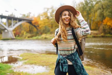 Ormanda şapka ve sırt çantası takan genç bir kadın, sonbaharda uzun ağaçların arasında huzurlu bir yürüyüş yapıyor. Orman banyosu. Etkin yaşam biçimi