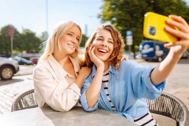 İki neşeli arkadaş bir kafe masasında oturmuş telefonla selfie çekiyorlar. Biri bir fincan kahve tutarken diğeri geniş bir gülümsemeyle anı kucaklıyor. Moda, güzellik, blog, turizm.