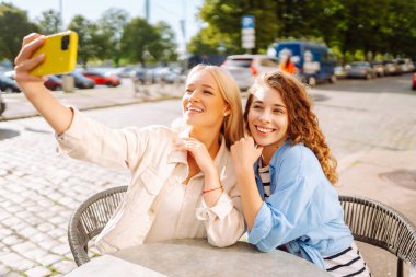 İki neşeli arkadaş bir kafe masasında oturmuş telefonla selfie çekiyorlar. Biri bir fincan kahve tutarken diğeri geniş bir gülümsemeyle anı kucaklıyor. Moda, güzellik, blog, turizm.