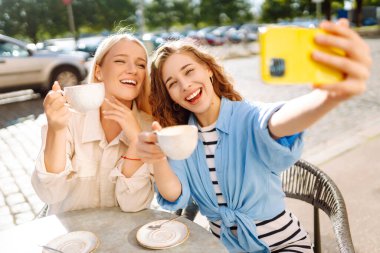 İki neşeli arkadaş bir kafe masasında oturmuş telefonla selfie çekiyorlar. Biri bir fincan kahve tutarken diğeri geniş bir gülümsemeyle anı kucaklıyor. Moda, güzellik, blog, turizm.