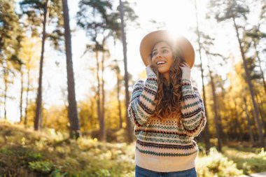 Parlak bir ormanda güneşli bir sonbahar gününün tadını çıkaran desenli süveter ve şapkalı neşeli bir kadın. Seyahat, turizm, hafta sonu.