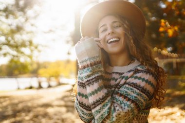Parlak bir ormanda güneşli bir sonbahar gününün tadını çıkaran desenli süveter ve şapkalı neşeli bir kadın. Seyahat, turizm, hafta sonu.