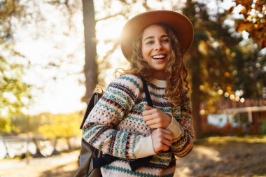 Parlak bir ormanda güneşli bir sonbahar gününün tadını çıkaran desenli süveter ve şapkalı neşeli bir kadın. Seyahat, turizm, hafta sonu.