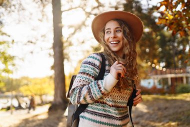 Parlak bir ormanda güneşli bir sonbahar gününün tadını çıkaran desenli süveter ve şapkalı neşeli bir kadın. Seyahat, turizm, hafta sonu.
