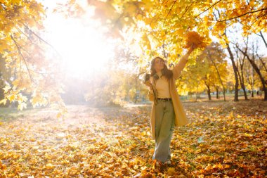 Parkta güneşli bir sonbahar gününün keyfini çıkaran neşeli bir kadın, canlı sarı yapraklar ve sakin bir atmosferle çevrili. Boş zaman aktivitesi.