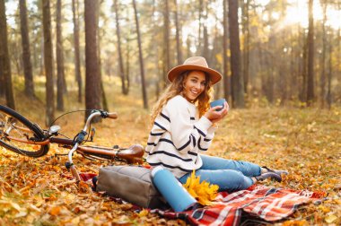 Genç bir kadın, renkli sonbahar yapraklarıyla çevrili bir battaniyenin üzerinde oturuyor. Sıcak bir içecek tutarken mutlu bir şekilde gülümsüyor. Yanında da sakin bir ormanda bisikleti var..