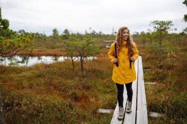 Sarı yağmurluklu neşeli genç bir kadın, tahta bir tahta kaldırımda duruyor, bir bataklığın sakin ortamını alırken gülümsüyor. Bataklıklar. Seyahat konsepti, keşif. 