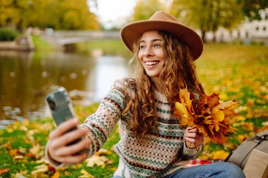 Nehir kenarında güneşli bir sonbahar gününün tadını çıkaran genç bir kadın renkli yapraklarla kaplı bir battaniyenin üzerinde otururken selfie çekiyor. Seyahat blogu. Macera, tatil konsepti.