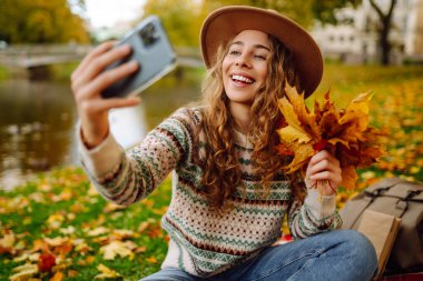 Nehir kenarında güneşli bir sonbahar gününün tadını çıkaran genç bir kadın renkli yapraklarla kaplı bir battaniyenin üzerinde otururken selfie çekiyor. Seyahat blogu. Macera, tatil konsepti.