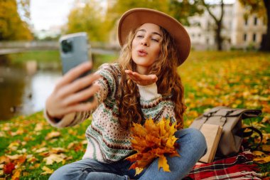 Nehir kenarında güneşli bir sonbahar gününün tadını çıkaran genç bir kadın renkli yapraklarla kaplı bir battaniyenin üzerinde otururken selfie çekiyor. Seyahat blogu. Macera, tatil konsepti.