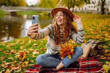 Nehir kenarında güneşli bir sonbahar gününün tadını çıkaran genç bir kadın renkli yapraklarla kaplı bir battaniyenin üzerinde otururken selfie çekiyor. Seyahat blogu. Macera, tatil konsepti.