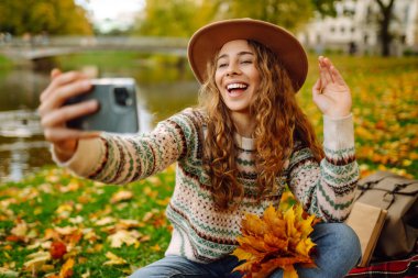 Nehir kenarında güneşli bir sonbahar gününün tadını çıkaran genç bir kadın renkli yapraklarla kaplı bir battaniyenin üzerinde otururken selfie çekiyor. Seyahat blogu. Macera, tatil konsepti.