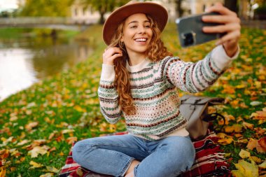 Nehir kenarında güneşli bir sonbahar gününün tadını çıkaran genç bir kadın renkli yapraklarla kaplı bir battaniyenin üzerinde otururken selfie çekiyor. Seyahat blogu. Macera, tatil konsepti.