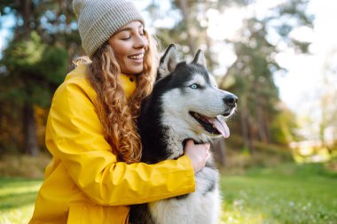 Sarı ceket giyen gülümseyen bir kadın, parktaki güneşli bir günün sıcaklığında güneşlenerek, canlı sonbahar yaprakları arasında boğuk köpeğiyle oynuyor. İri köpek. Dostluk, evcil hayvan ve insan.