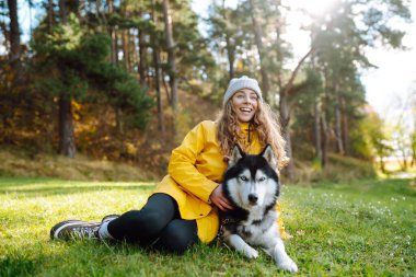 Sarı ceket giyen gülümseyen bir kadın, parktaki güneşli bir günün sıcaklığında güneşlenerek, canlı sonbahar yaprakları arasında boğuk köpeğiyle oynuyor. İri köpek. Dostluk, evcil hayvan ve insan.