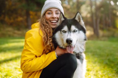 Sarı ceket giyen gülümseyen bir kadın, parktaki güneşli bir günün sıcaklığında güneşlenerek, canlı sonbahar yaprakları arasında boğuk köpeğiyle oynuyor. İri köpek. Dostluk, evcil hayvan ve insan.