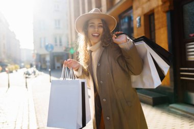 Genç bir kadın, güzel bir günde alışveriş çılgınlığının tadını çıkarırken, canlı bir şehir ortamında birkaç alışveriş poşetini tutarken parlak bir şekilde gülümsüyor. Tüketim, satış, alım, alışveriş, yaşam tarzı konsepti.