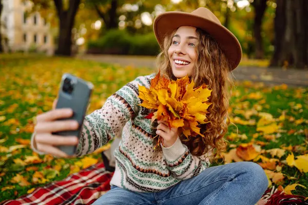 Nehir kenarında güneşli bir sonbahar gününün tadını çıkaran genç bir kadın renkli yapraklarla kaplı bir battaniyenin üzerinde otururken selfie çekiyor. Seyahat blogu. Macera, tatil konsepti.