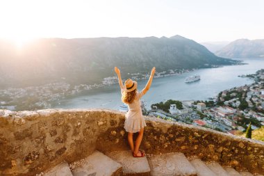 Karadağ 'ın Kotor kentindeki manzaralı bir manzarada gün batımını kutlayan bir kadın, kollarını körfez ve dağların üzerine kaldırdı. Turizm, rahatlama, gençlik ve doğa kavramı.