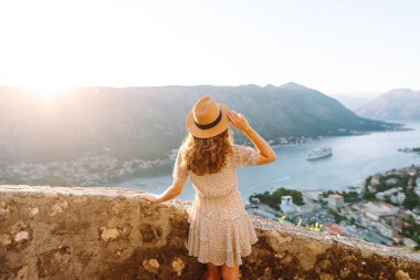 Karadağ 'ın Kotor kentindeki manzaralı bir manzarada gün batımını kutlayan bir kadın, kollarını körfez ve dağların üzerine kaldırdı. Turizm, rahatlama, gençlik ve doğa kavramı.