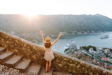 Karadağ 'ın Kotor kentindeki manzaralı bir manzarada gün batımını kutlayan bir kadın, kollarını körfez ve dağların üzerine kaldırdı. Turizm, rahatlama, gençlik ve doğa kavramı.