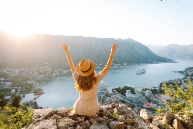 Karadağ 'ın Kotor kentindeki manzaralı bir manzarada gün batımını kutlayan bir kadın, kollarını körfez ve dağların üzerine kaldırdı. Turizm, rahatlama, gençlik ve doğa kavramı.