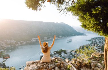 Karadağ 'ın Kotor kentindeki manzaralı bir manzarada gün batımını kutlayan bir kadın, kollarını körfez ve dağların üzerine kaldırdı. Turizm, rahatlama, gençlik ve doğa kavramı.