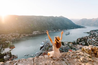 Karadağ 'ın Kotor kentindeki manzaralı bir manzarada gün batımını kutlayan bir kadın, kollarını körfez ve dağların üzerine kaldırdı. Turizm, rahatlama, gençlik ve doğa kavramı.