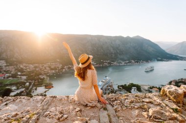 Karadağ 'ın Kotor kentindeki manzaralı bir manzarada gün batımını kutlayan bir kadın, kollarını körfez ve dağların üzerine kaldırdı. Turizm, rahatlama, gençlik ve doğa kavramı.