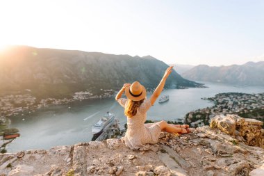 Karadağ 'ın Kotor kentindeki manzaralı bir manzarada gün batımını kutlayan bir kadın, kollarını körfez ve dağların üzerine kaldırdı. Turizm, rahatlama, gençlik ve doğa kavramı.