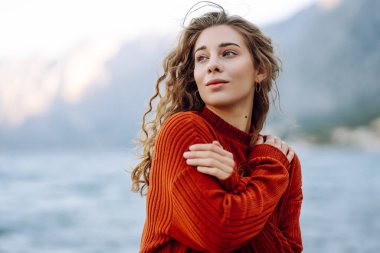 Genç bir kadın, gölün kenarında güneşli bir öğleden sonra, arkasında dağlar olan rahat bir süveterin tadını çıkarıyor. Moda tarzı. Yaşam tarzı, seyahat, turizm.